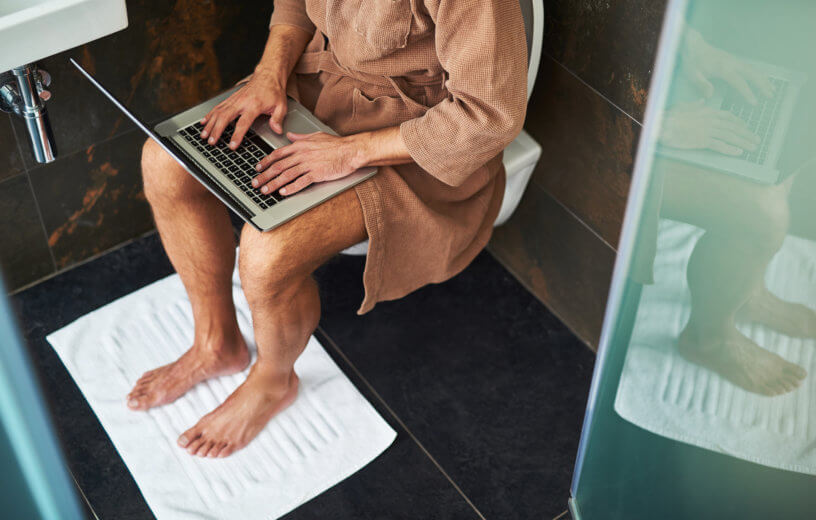Man in bathrobe using laptop while sitting on toilet at home