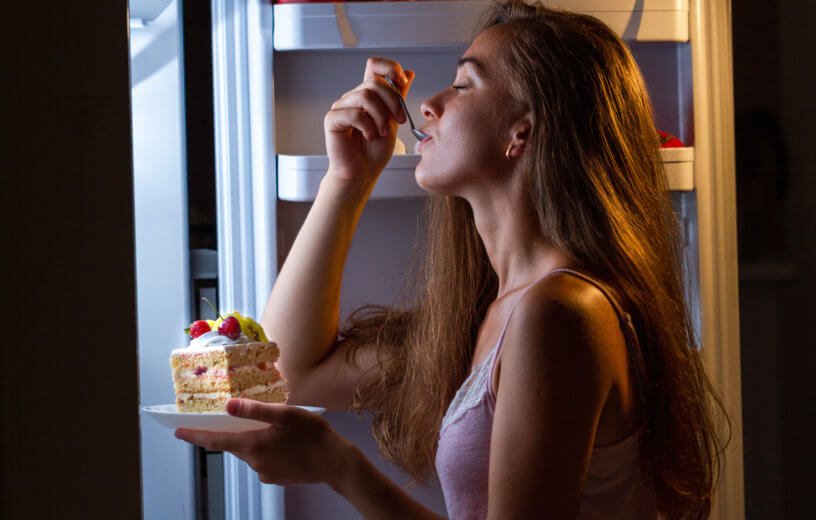 Woman enjoying late night snacking, eating cake in front of refrigerator