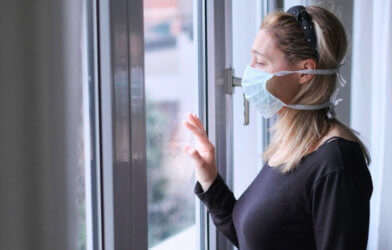 Woman wearing mask looking out window while under quarantine isolation for coronavirus / COVID-19 outbreak