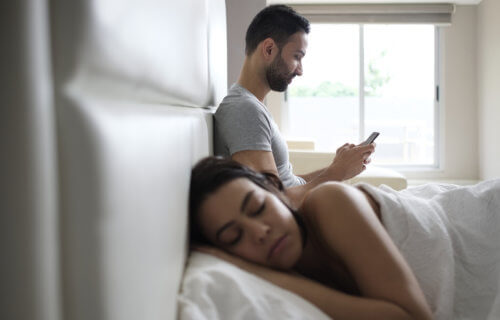 Man texting in bed on smartphone next to sleeping wife